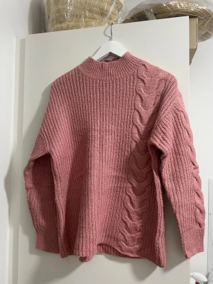Rosa stickad tröja med flätmönster - Mysig rosa stickad tröja med höghals och ett snyggt flätmönster på ena sidan framtill. Tröjan har lång ärm och är ribbstickad för extra struktur. Perfekt för kyliga dagar och enkel att styla med jeans eller kjol.