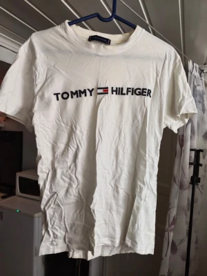 Vit Tommy  t-shirt Helg REA NU! - Snygg vit t-shirt från Tommy  med klassisk logga tryckt framtill i svart, rött och blått. T-shirten har rund halsringning och korta ärmar. Tillverkad i mjuk bomull för en skön känsla och enkel stil. Kan finnas någon fläck men kan få bort .