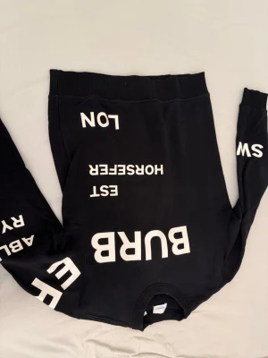 Svart sweatshirt från Burberry - Cool svart sweatshirt från Burberry med vita tryckta bokstäver och logotexter över hela tröjan. Oversized känsla och ribbade muddar vid ärmslut och midja. Perfekt för dig som gillar streetwear och vill sticka ut med en unik design.