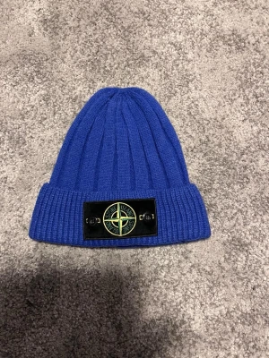 Stone Island beanie!! - Snygg blå ribbstickad mössa från Stone Island med uppvikt kant och den klassiska logotypen framtill på ett svart märke. Tillverkad i mjukt material som håller dig varm och ger en clean streetwear-look.