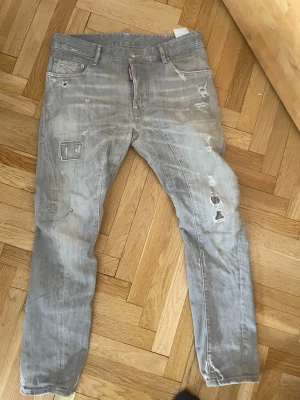 Grå slitna jeans från Dsquared2 - Säljer ett par grå jeans från Dsquared2 med distressed detaljer och patchade partier. Jeansen har raka ben, fem fickor och synliga slitningar för en cool, avslappnad look. Perfekta för dig som gillar streetwear och en trendig stil. Sitter som W 30
