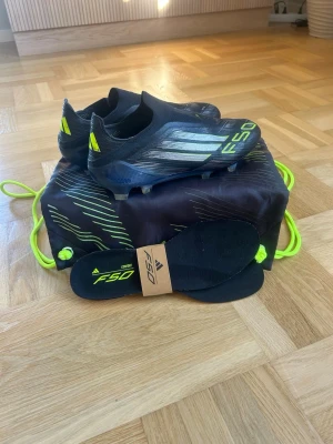 Adidas F50 Svarta Fotbollsskor - Säljer ett par svarta Adidas F50 fotbollsskor med neongula detaljer och silvriga ränder. Mycket bra skick och är bara använda ett fåtal gånger. Du får med skor, skopåse och extra sulor. Köpta från Unisport
