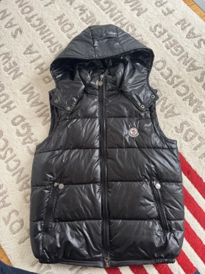 Svart dunväst från Moncler med luva - Snygg svart dunväst från Moncler med avtagbar huva och två dragkedjeförsedda fickor. Västen har ett glansigt yttertyg och klassisk Moncler-logga på bröstet. Perfekt för lager på lager och streetwear-vibe.