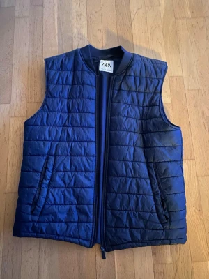Mörkblå dunväst från ZARA - Snygg mörkblå dunväst från ZARA med quiltat mönster och dragkedja framtill. Västen har två fickor med dragkedja och ribbad krage för extra stilpoäng. Passar M och L. Inte använd över 3 gånger.