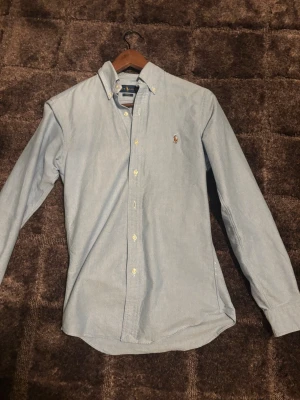 Ljusblå skjorta från Ralph Lauren - Klassisk ljusblå skjorta från Ralph Lauren med button-down krage och broderad logga på bröstet. Skjortan har långa ärmar, vita knappar och är tillverkad i mjuk bomull. Perfekt för en clean och stilren look.