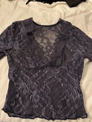 Mörklila spetsblus med v-ringning - Snygg mörklila blus i transparent spets med djup v-ringning och trekvartsärm. Blusen har ett blommigt spetsmönster som ger en romantisk vibe och passar perfekt till lager på lager eller som statement-plagg.