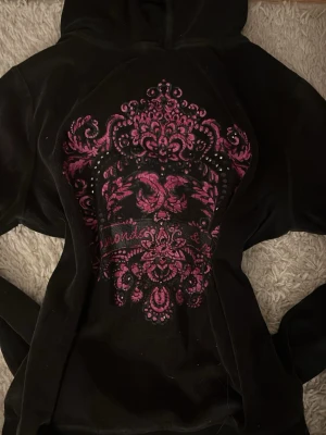 Svart hoodie från Hunkemöller S - Mysig svart hoodie från Hunkemöller med rosa dekorativt tryck och små glittriga stenar på bröstet. Tröjan har huva och är tillverkad i mjukt material, perfekt för chill dagar hemma. Snygg detalj med fåglar och mönster framtill.