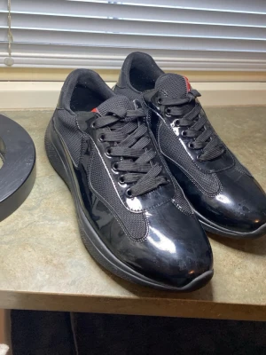 Svarta sneakers från Prada - Snygga svarta sneakers från Prada med glansig finish och meshdetaljer. Skorna har svarta skosnören, diskret röd logga på plösen och en chunky sula. Perfekta för dig som gillar en clean och modern look.