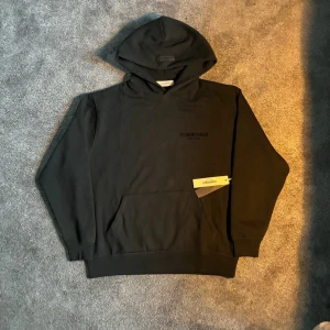 Svart Essentials, hoodie - Snygg svart hoodie från Essentials hoodid med diskret logga på bröstet och stor logga på ryggen. Klassisk passform med huva och ribbade muddar. Tillverkad i mjukt bomullsmaterial som känns riktigt skönt mot huden. Perfekt för en clean och stilren streetwear-look.