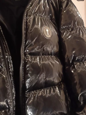 Svart pufferjacka från Trapstar - Säljer en svart, glansig pufferjacka från Trapstar med tydlig logga på bröstet och cool Trapstar-tryck på ryggen. Jackan har dragkedja framtill och breda, vadderade paneler som ger en riktigt fet streetwear-look. Perfekt för kalla vinterdagar.