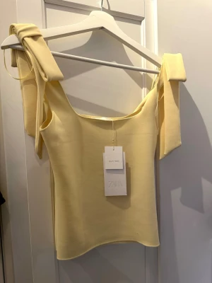 Gul topp med knytband från Zara - Supersöt ljusgul topp från Zara med breda axelband som knyts till rosetter. Toppen har en rak, croppad passform och är tillverkad i ett mjukt, stretchigt material som känns bekvämt mot huden. Perfekt för att addera färg till din outfit!