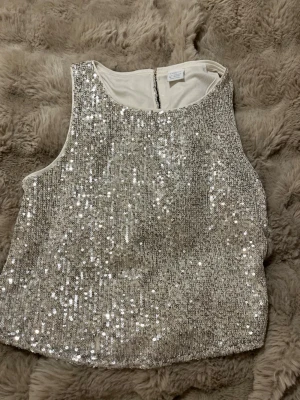 Glittrig silver/guld paljetlinne från Lindex - Supersnygg ärmlös topp från Lindex i silver/guld med massor av paljetter som ger ett riktigt glittrigt intryck. Toppen har rund halsringning och en kanpp i ryggen. Perfekt för dig som vill sticka ut och glänsa lite extra.