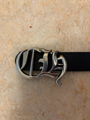 Chrome Hearts Svart bälte - Snyggt svart bälte med ett stort, silvrigt spänne i unik form och flera dekorativa metalldetaljer längs remmen. Perfekt accessoar för att lyfta din outfit med en edgy touch. Passar till jeans eller kjol för en cool stil. Chrome hearts