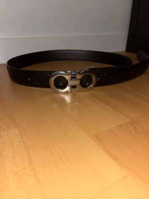 Svart skärp från Salvatore Ferragamo - Snyggt svart skärp från Salvatore Ferragamo med ikoniskt silverfärgat spänne och präglat mönster längs hela remmen. Ända defekt är hålet som man ser på sista bilden Perfekt accessoar för att lyfta vilken outfit som helst med en lyxig touch.