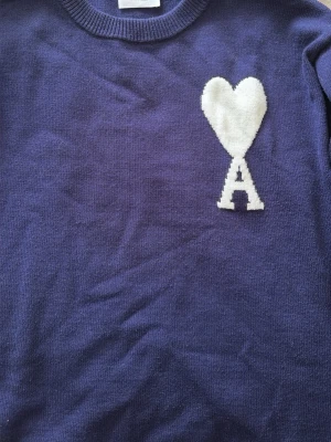 Mörk Blå stickad tröja från Ami/alexandre mattussi - Säljer en mörk blå stickad tröja från Ami med ett vitt hjärta och bokstaven A på bröstet. Tröjan har rund halsringning och är tillverkad i mjukt stickat material. Perfekt för dig som gillar stilrena detaljer och vill ha något unikt i garderoben.