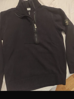 Svart stickad tröja Stone Island - Säljer en svart stickad tröja Stone Island med halv dragkedja framtill och hög krage. Tröjan har ribbade muddar och nederkant samt den klassiska Stone Island-loggan på vänster ärm. Perfekt för dig som gillar streetwear och snygga detaljer.