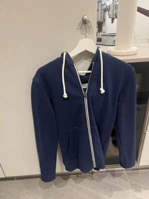Mörkblå hoodie med dragkedja från pier one - Mörkblå hoodie från pier one med vit dragkedja och vita snören i huvan. Klassisk modell med känguruficka framtill och mjukt, tjockt material som känns riktigt skönt. Perfekt för chill dagar eller när du vill ha en enkel och stilren look.