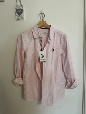 Rosa randig skjorta från U.S. Polo Assn. - Säljer en klassisk rosa och vit randig skjorta från U.S. Polo Assn. Skjortan har lång ärm, knappar framtill och broderad logga på bröstet. Perfekt för en clean och stilren look.