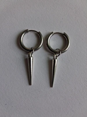 Silvriga hoops med spetsiga hängen - Säljer ett par silvriga örhängen i form av hoops med avlånga, spetsiga hängen. Örhängena har en minimalistisk och edgy design som ger en cool touch till din stil. Materialet ser ut att vara metall och de har en blank finish.