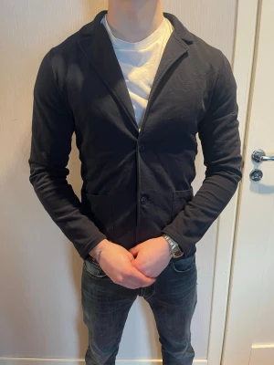 Kavajtröja från Woolrich med knappar - Snygg svart kavajtröja med två knappar framtill och två fickor. Tröjan har en klassisk krage och är tillverkad i ett mjukt, stretchigt material. Perfekt för en stilren och avslappnad look.