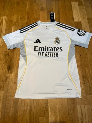 Real Madrid home kit 25/26 X Bellingham 5 - Vit Real Madrid matchtröja från Adidas med svarta detaljer, gula linjer och klubbmärke på bröstet. Tryck med 'Emirates Fly Better' på framsidan och 'Bellingham 5' på ryggen. Tillverkad i lätt funktionsmaterial Aeroready, perfekt för fotbollsträning.