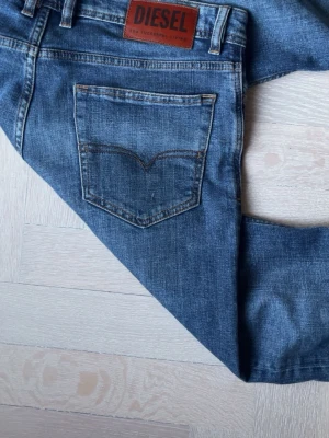 Blå jeans från Diesel - Säljer ett par klassiska blå jeans från Diesel med fem fickor och snygga slitningar. Jeansen har en rak passform och detaljerade sömmar på bakfickan. Tillverkade i slitstark denim med läderpatch bak i midjan.