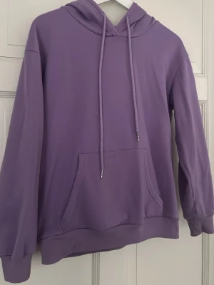 Lila hoodie med magficka och huva - Snygg lila hoodie med stor magficka och justerbar huva med långa snören. Hoodien har ribbade muddar vid ärmslut och nederkant. Perfekt för en avslappnad och trendig stil. Tillverkad i mjukt bomullsliknande material.