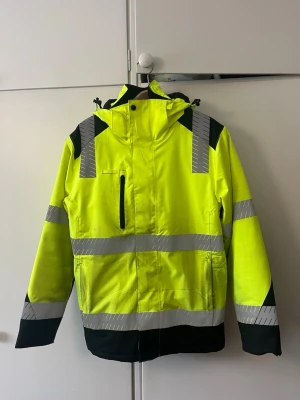 Gul vindjacka med reflexdetaljer - Snygg gul vindjacka från Texstar med svarta detaljer och flera reflexband för extra synlighet. Jackan har huva, dragkedja framtill, bröstficka med dragkedja och justerbara muddar. Perfekt för dig som vill sticka ut och hålla dig synlig i alla väder. Behövs tvättas ( storlek xs men skulle säga s) 