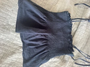 Svart linnetopp med knytband - Säljer en svart linnetopp med smockad överdel och tunna axelband som knyts upptill. Toppen har en luftig och flowy passform med volangkant upptill. Perfekt för varma dagar och enkel att styla till olika looks.