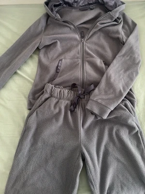 Grått fleece loungewear-set med huva - Mysigt grått loungewear-set i fleece med dragkedja, huva och fickor på tröjan samt matchande byxor med resår och satinband i midjan. Perfekt för chill dagar hemma. Materialet är mjukt och värmande.