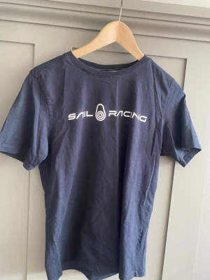 Marinblå Sail Racing t-shirt - Snygg marinblå t-shirt från Sail Racing med vit logga tryckt på bröstet och en liten logga i nacken. Klassisk passform och rund halsringning. Tillverkad i mjuk bomull som känns skön mot huden. Perfekt för en avslappnad och sportig stil.