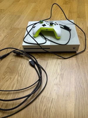 Xbox One S med vit och grön kontroll - Säljer en Xbox One S i vit färg med tillhörande vit och grön handkontroll. Konsolen har en stilren, rektangulär design med ventilerad ovansida och medföljande kablar. Perfekt för gaming och streaming hemma.