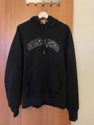 Svart hoodie Namasenda merch - Snygg svart hoodie från Namasenda med huva och dragsnören. Framtill står det 'NAMA★SENDA' i glittriga stenar som ger en cool vibe. Hoodien har en stor magficka och ribbade muddar vid ärmar och nederkant. 