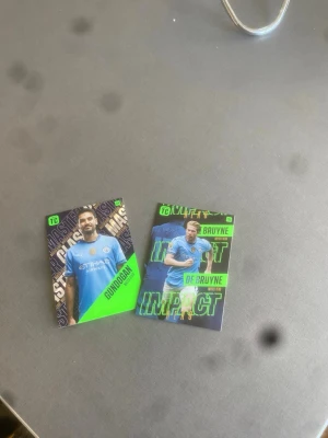 Fotbollssamlarbilder Manchester City - Två samlarkort med fotbollsspelare från Manchester City. Kortens design går i blått, grönt och svart med spelarnas namn och klubbtröja tydligt synliga. Perfekt för dig som gillar fotboll och vill utöka din samling.