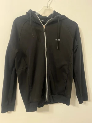 Svart zip hoodie från BOSS - Säljer en svart hoodie från BOSS med dragkedja framtill och justerbar huva. Diskret BOSS-logga på bröstet och större logga på ryggen. Tillverkad i mjukt bomullsmaterial och har två fickor på sidorna. Perfekt för en avslappnad och stilren look.