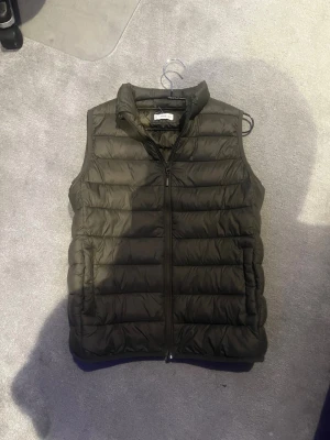 Grön dunväst med dragkedja - Snygg grön dunväst med quiltad design och hög krage. Västen har dragkedja framtill och två sidofickor. Perfekt att slänga över en hoodie eller tröja för extra värme. Lätt och smidig att bära, passar till många olika stilar.