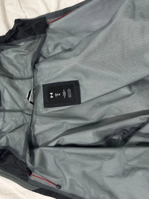 Svart vindjacka från Under Armour - Snygg svart vindjacka från Under Armour med röd logga på framsidan. Jackan har meshfoder på insidan för extra ventilation och dragkedja med röda detaljer. Perfekt för blåsiga dagar och sportiga outfits.