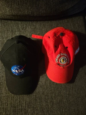 NASA och Chicago Blackhawks kepsar - Två snygga kepsar: en svart med klassisk NASA-logga broderad framtill och en röd Chicago Blackhawks-keps med broderad hockeylogga. Båda har böjd skärm och justerbar rem bak. Perfekta för dig som gillar sport eller rymd! Materialet är bomull.
