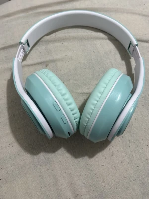 Mintgröna trådlösa over-ear hörlurar - Snygga mintgröna over-ear hörlurar med vita detaljer. De har vadderade öronkuddar och justerbart huvudband för extra komfort. Utrustade med knappar på sidan för enkel styrning av musik och samtal. Perfekta för dig som vill ha stilrena och bekväma hörlurar.