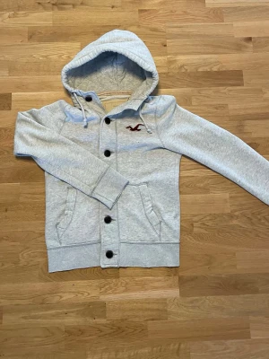 Ljusgrå hoodie från Hollister - Ljusgrå hoodie från Hollister med knappar framtill och broderad vinröd logga på bröstet. Hoodien har dragsko i huvan och två fickor fram. Tillverkad i mjukt sweatshirtmaterial som är perfekt för en avslappnad stil.
