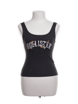 Svart linnetopp från Hollister - Snygg svart linnetopp från Hollister med broderad logga och små blommor framtill. Toppen har breda axelband och en figurnära passform som ger en clean och trendig look. Perfekt att styla med jeans eller kjol för en chill vibe.