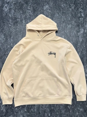 Beige hoodie från Stüssy med tryck - Säljer en beige hoodie från Stüssy med svart logga på bröstet och stort tryck med blå tärningar och logga på ryggen. Hoodien har huva, känguruficka och ribbade muddar. Materialet känns mjukt och skönt, perfekt för en chill streetwear-look.