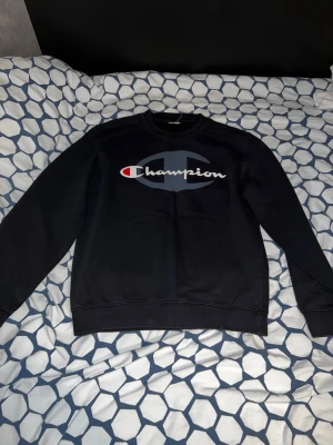 Svart Champion sweatshirt - Säljer en svart sweatshirt från Champion med stort logotryck i vitt, blått och rött på bröstet. Tröjan har rund halsringning och ribbade muddar vid ärmslut och nederkant. Perfekt för en avslappnad och sportig stil.