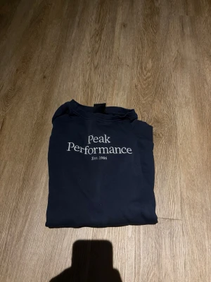 Mörkblå sweatshirt Peak Performance - Mörkblå sweatshirt från Peak Performance med vit logga och text 'Est. 1986' på bröstet. Klassisk rund halsringning och långa ärmar. Perfekt för en avslappnad och stilren look.