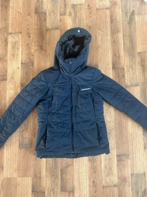 Mörkblå dunjacka Peak Performance - Snygg mörkblå dunjacka från Peak Performance med huva och dragkedja framtill. Jackan har en bröstficka med dragkedja och diskret logga på vänster sida. Litet hål på höger armen som knappt syns. M men liten i storlek 