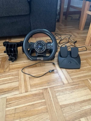 Ratt och pedaler till spelkonsol - Säljer ett svart racing ratt- och pedalset från Nacon, perfekt för bilspel på konsol eller PC. Ratten har flera knappar och en klassisk design med gummigrepp. Pedalerna är i plast och kopplas in med USB. Fäste för bord medföljer.