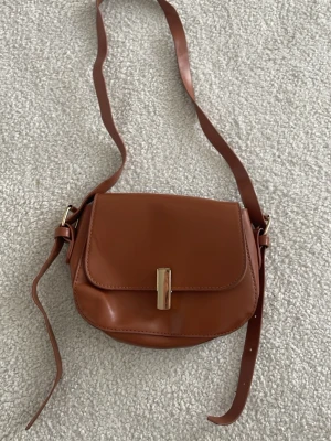 Brun axelväska i läderlook - Snygg brun axelväska i läderimitation med justerbart axelband och guldfärgat spänne framtill. Väskan har rundad form och stilren design som passar till många outfits. Perfekt storlek för det viktigaste och enkel att bära crossbody. Köpt på björk&frihet secondhand. Prislapp finns kvar i väskan. Har inte använt den jag köpte den. Säljer för så mycket jag köpte den för men kan diskutera pris 