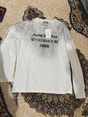 Vit långärmad topp Acne Studios - Vit långärmad topp från Acne Studios med tryck 'STOCKHOLM 1996' i svart framtill. Tunn och mjuk bomullskvalitet med rund halsringning. Perfekt för en clean och stilren look.