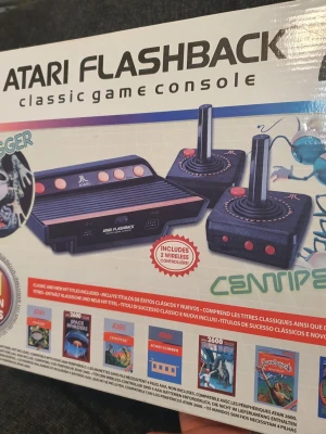 Atari Flashback spelkonsol - Retro. Atari Flashback spelkonsol med svart och brun design. Konsolen har klassisk rektangulär form och kommer med två trådlösa joystick-kontroller. Perfekt för dig som gillar retrospel och vill återuppleva gamla klassiker.
