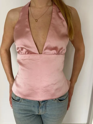 Rosa satin halterneck topp med knyte - Snygg ljusrosa halterneck topp i satin med djup v-ringning och öppen rygg. Toppen knyts i ryggen med band för en justerbar passform. Perfekt att styla med jeans eller kjol för en snygg look. 💓